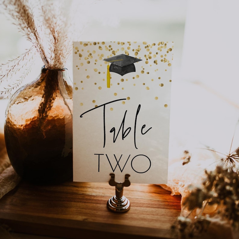 K&ouml;nnte beinhalten: Eine wei&szlig;e Karte mit goldenem Glitzer und einer Illustration einer schwarzen Graduation Cap. Die Karte tr&auml;gt die Aufschrift "Tisch Zwei" in schwarzer Schrift.