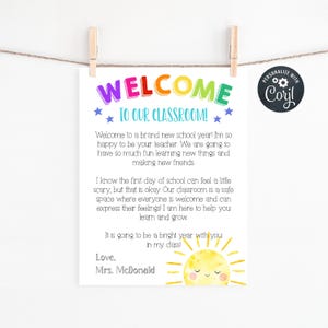 May include: A white classroom welcome sign featuring the colorful text WELCOME TO OUR CLASSROOM! with blue stars, a message from Mrs. McDonald, and a yellow watercolor sun illustration at the bottom right corner, suspended by two wooden clips.