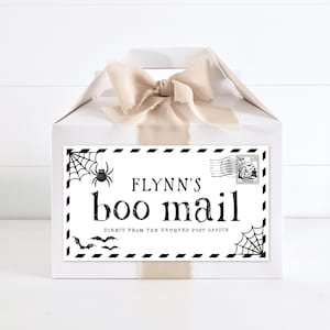May include: A white gift box with a beige ribbon bow on top, featuring a rectangular label that reads FLYNN'S boo mail DIRECT FROM THE HAUNTED POST OFFICE, decorated with black spiderwebs, a spider, bats, and a postage stamp with pumpkins.