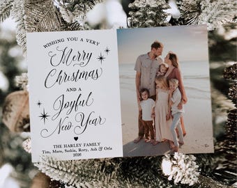 May include: A rectangular holiday card featuring a white left panel with black calligraphy reading Wishing You A Very Merry Christmas And A Joyful New Year, The Harley Family, and 2026. The right panel shows a family of six on a beach.