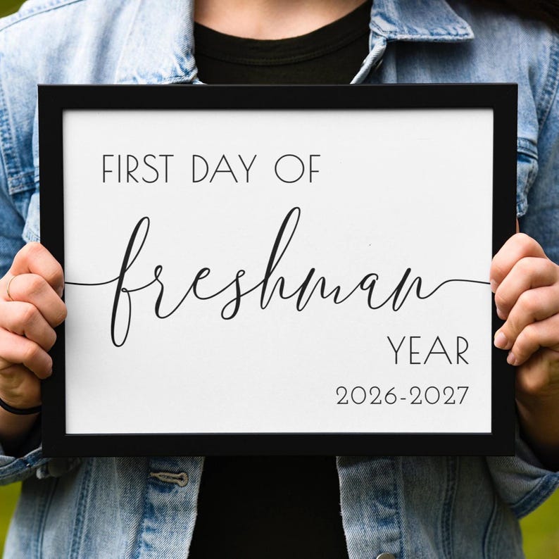 Op de afbeelding: Een zwart omrand bord met de tekst "FIRST DAY OF freshman YEAR 2026-2027" in een mix van sans-serif en cursieve lettertypen. Het bord wordt vastgehouden door een persoon die een spijkerjasje draagt.