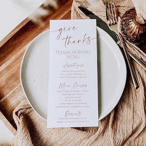 May include: A white rectangular Thanksgiving menu card with copper-colored text reading Give Thanks, Thanksgiving Menu, Appetizers, Main Courses, and Desserts, placed on a round white plate with a gold fork and knife beside it.