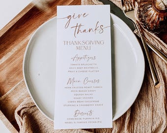 May include: A white rectangular Thanksgiving menu card with copper-colored text reading Give Thanks, Thanksgiving Menu, Appetizers, Main Courses, and Desserts, placed on a round white plate with a gold fork and knife beside it.