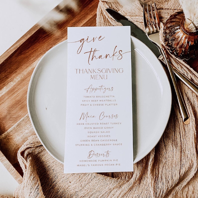 May include: A white rectangular Thanksgiving menu card with copper-colored text reading Give Thanks, Thanksgiving Menu, Appetizers, Main Courses, and Desserts, placed on a round white plate with a gold fork and knife beside it.