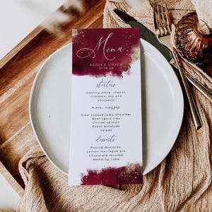 May include: A wedding menu card with a burgundy and gold watercolor design. The menu lists starters, main courses, and desserts. The names Alicia & Steven and the date 01.16.23 are at the top. The card is on a white plate with silverware.