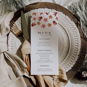 May include: A white menu card with a fall leaf design, placed on a layered plate setting with a gold fork and knife. The menu lists appetizers, main courses, and desserts, including pumpkin pie.