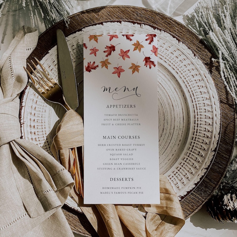 May include: A white menu card with a fall leaf design, placed on a layered plate setting with a gold fork and knife. The menu lists appetizers, main courses, and desserts, including pumpkin pie.