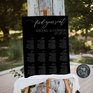 Peut inclure: Un panneau de plan de table de mariage rectangulaire noir avec du texte blanc indiquant Find your seat, Rachel & Connor, 07.07.24, et listant les noms des invités sous douze tables numérotées. Le panneau est posé sur un chevalet en bois.