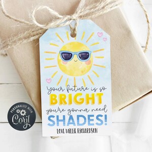 May include: A rectangular gift tag with a light blue background featuring a yellow sun wearing blue sunglasses. The text reads: Your future is so BRIGHT you're gonna need SHADES! Love Millie Richardson. The tag is tied with twine to a brown package.