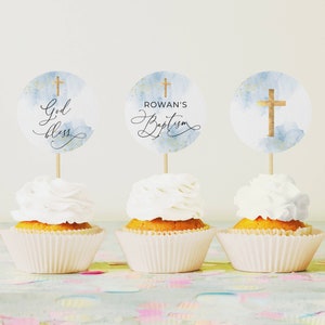 May include: Three cupcakes with white frosting topped with round blue and gold watercolour cupcake toppers. The toppers feature a gold cross and the text "God Bless", "Rowan's Baptism", and a gold cross.