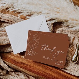 May include: A rectangular thank you card with a terracotta background, featuring a white line-art floral illustration on the left. The text reads thank you, rosie & wyatt, and 07.07.24 in white. A white envelope is positioned behind the card.