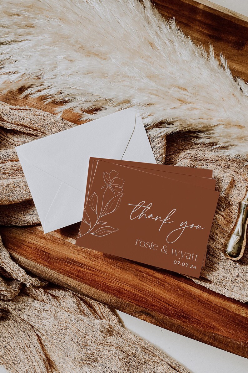 May include: A rectangular thank you card with a terracotta background, featuring a white line-art floral illustration on the left. The text reads thank you, rosie & wyatt, and 07.07.24 in white. A white envelope is positioned behind the card.