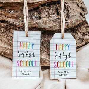 May include: Two white gift tags with a lined background and rainbow lettering that reads "HAPPY first day of SCHOOL!" with the sender's name below. The tags are hanging from twine and are against a rustic wooden background.