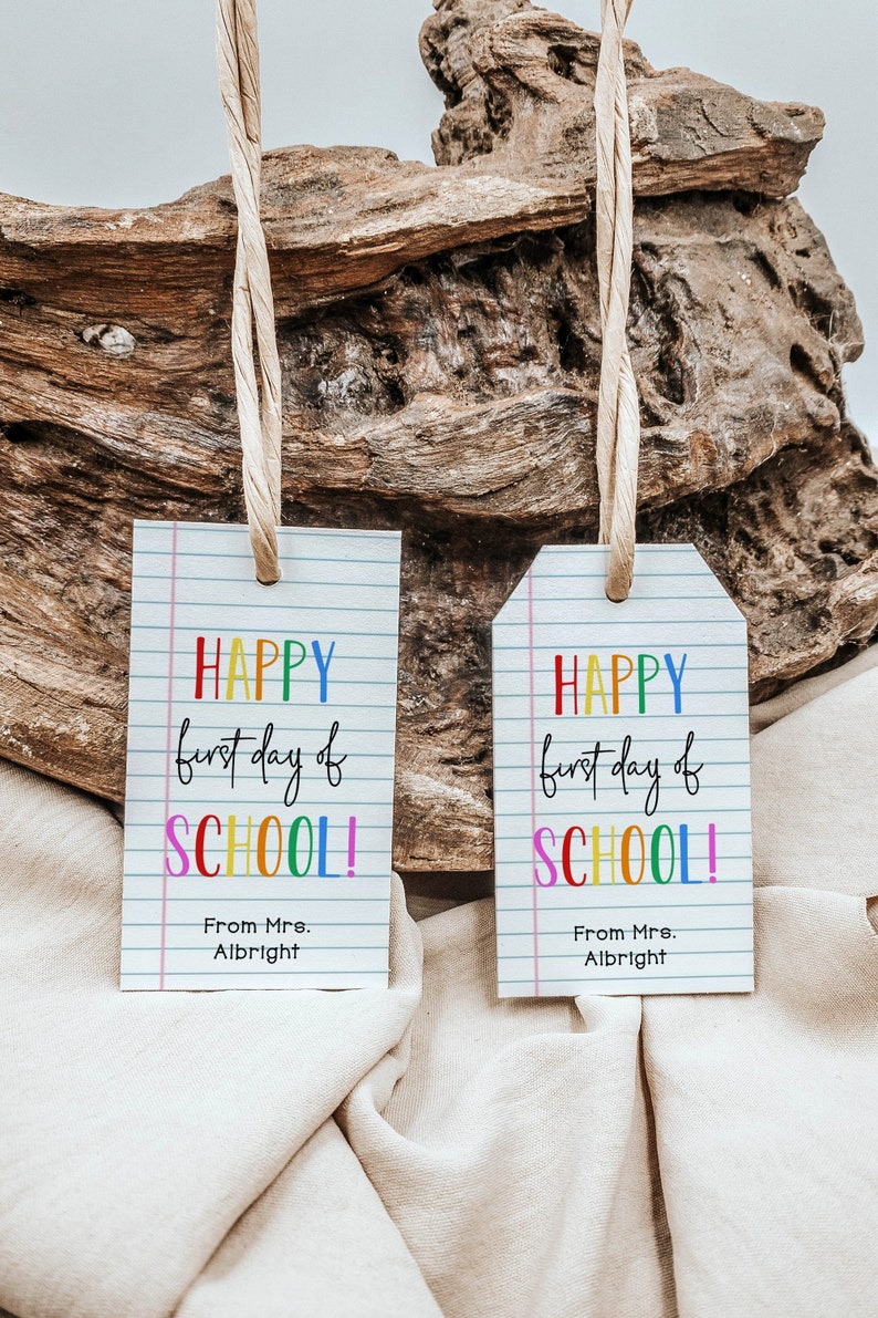 May include: Two white gift tags with a lined background and rainbow lettering that reads "HAPPY first day of SCHOOL!" with the sender's name below. The tags are hanging from twine and are against a rustic wooden background.