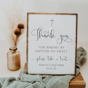 以下が含まれることがあります： 金色の枠と上部に黒い十字架が付いた白い看板。看板には「私の洗礼をこんなに甘くしてくれたことに感謝します。どうぞ、おやつをどうぞ。マエヴの洗礼 10.10.25」と書かれています。