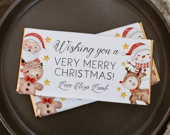 May include: White chocolate bar wrapper with a festive Christmas design. The wrapper features illustrations of Santa Claus, a gingerbread person, a snowman, and a reindeer. The text on the wrapper reads "Wishing you a very merry Christmas! Love Eliza Lamb."