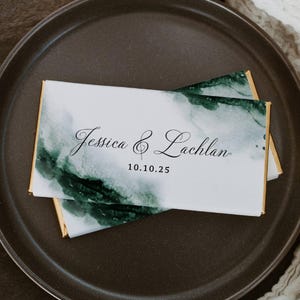 Puede incluir: Una barra de chocolate blanco con un diseño de acuarela verde y el texto "Jessica & Lachlan" y la fecha "10.10.25".