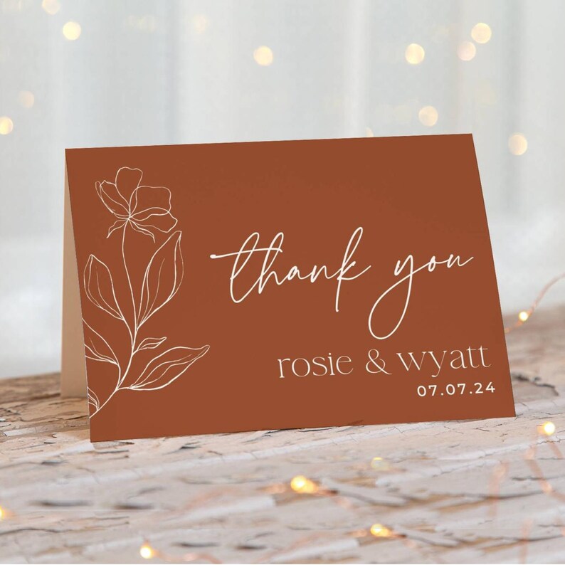 May include: A rectangular folded card with a terracotta background featuring a white line art floral illustration on the left. The text reads thank you in a script font, followed by the names rosie & wyatt and the date 07.07.24 in white.