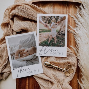 May include: Two white photo cards with black borders and text. The top card shows a baby girl standing in a grassy field. The text reads "Eleven Months". The bottom card shows a baby lying on a white blanket. The text reads "Three Months".