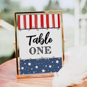 Op de afbeelding: Een ingelijst tafelschild met een patriottisch thema. De bovenkant heeft rood-witte strepen, de onderkant heeft een blauwe achtergrond met witte sterren en de tekst luidt "Table ONE" in zwart script en blokletters.
