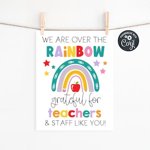 May include: A white print with the text "WE ARE OVER THE RAINBOW grateful for teachers & STAFF LIKE YOU!" A rainbow, apple, and stars are illustrated on the print. The print is hanging from a clothesline with wooden clothespins.