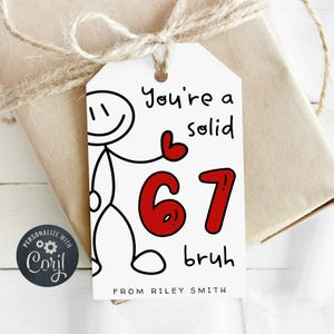 Puede incluir: Una etiqueta de regalo blanca con un hombre de palo y el texto "You're a solid 67 bruh". El número 67 es rojo. La etiqueta está atada a un regalo envuelto con hilo.