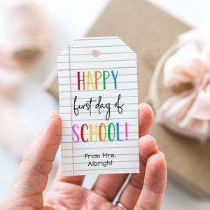 May include: A colorful gift tag with the text "HAPPY first day of SCHOOL!" and "From Mrs. Albright" on a white background with pink lines.