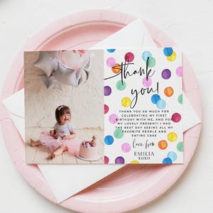 Può includere: Un biglietto di ringraziamento con la foto di una bambina e il testo "Grazie!" e "Grazie mille per aver festeggiato il mio primo compleanno con me..." Il biglietto ha pois colorati ed è su un piatto rosa.