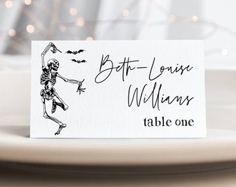 Op de afbeelding: Een wit placecard met een zwart skelet dat danst en twee vleermuizen die erboven vliegen. De tekst op de kaart luidt "Beth-Louise Williams tafel één".