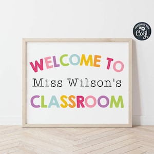 May include: A rectangular sign in a light wood frame featuring the text WELCOME TO Miss Wilson's CLASSROOM. The words WELCOME TO and CLASSROOM are printed in large, multi-coloured sans-serif letters, while Miss Wilson's is in black text.
