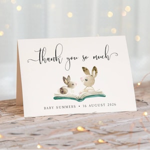 May include: A folded white card featuring the black cursive text thank you so much at the top. Below, a watercolour illustration shows two brown rabbits sitting on an open green book. The text BABY SUMMERS 16 AUGUST 2026 is printed at the bottom.