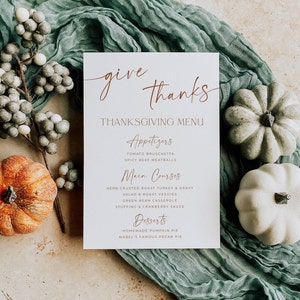 May include: A white rectangular Thanksgiving menu card with copper-colored text reading give thanks, Appetizers, Main Courses, and Desserts, surrounded by a green fabric runner, a branch of grey berries, one orange pumpkin, and two white pumpkins.