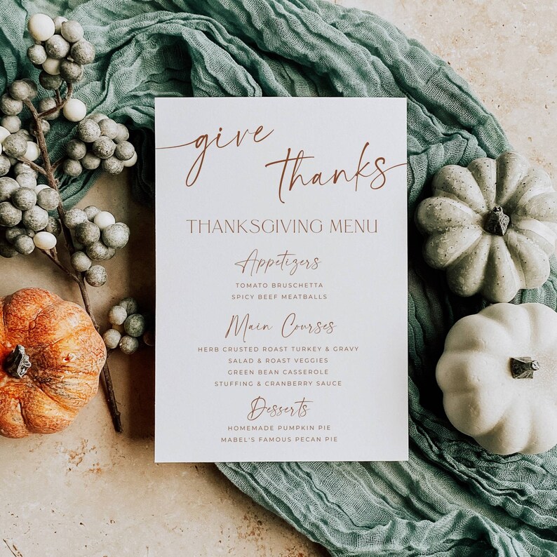 May include: A white rectangular Thanksgiving menu card with copper-colored text reading give thanks, Appetizers, Main Courses, and Desserts, surrounded by a green fabric runner, a branch of grey berries, one orange pumpkin, and two white pumpkins.