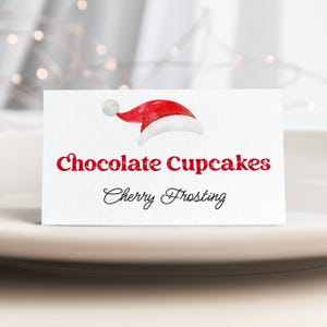Może przedstawiać: Biała karta z akwarelową ilustracją czapki Świętego Mikołaja. Na karcie widnieje napis "Chocolate Cupcakes" czerwoną czcionką i "Cherry Frosting" czarną kursywą. Karta leży na białym talerzu.