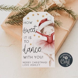 以下が含まれることがあります： サンタ帽と赤いチュチュを着たバレリーナのイラストが描かれたクリスマスギフトタグ。「How sweet it is to dance with you! Merry Christmas! Love Ashley.」と書かれています。金色の星がタグを飾っています。