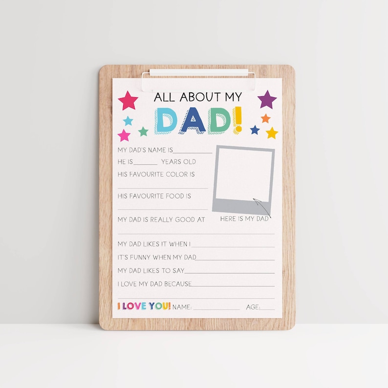 May include: A wooden clipboard holding a white questionnaire titled ALL ABOUT MY DAD. The sheet features colorful stars, blank lines for writing, and a gray square frame labeled HERE IS MY DAD with an arrow pointing to it. The bottom reads I LOVE YOU!