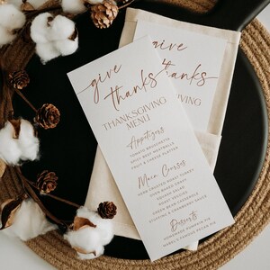 May include: Two white paper Thanksgiving menu cards with copper-colored text reading "give thanks" and listing courses including appetizers, main courses, and desserts. The cards are placed on a cream-colored fabric napkin.