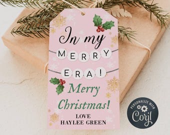 May include: A pink Christmas gift tag with the text "In my MERRY ERA! Merry Christmas!" and the name "HAYLEE GREEN". The tag has holly and snowflake designs, and is attached to a wrapped gift with greenery.