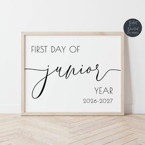 May include: A framed print with the text "FIRST DAY OF junior YEAR 2026-2027" in black script and sans-serif fonts. The frame is a light wood color, and the background is white. A small circular logo in the upper right corner reads "Instant Download DIY Print."