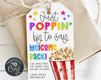 May include: A gift tag with the text "Just POPPIN' by to say welcome back!" in colorful letters. The tag features a cartoon popcorn bucket and is tied to a wrapped gift with twine.