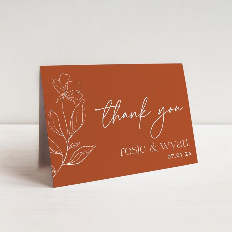 May include: A folded thank you card with a burnt orange background featuring a white line art floral illustration on the left side. The text reads thank you in a script font, followed by the names rosie & wyatt and the date 07.07.24.