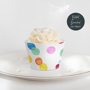 Op de afbeelding: Een witte cupcake met witte frosting en sprinkles, in een cupcake-verpakking met een kleurrijk aquarel-polka-dot-patroon. De verpakking heeft een witte achtergrond met meerkleurige stippen.