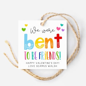 May include: A white square gift tag with a colorful message and a twine string. The text reads "We were bent to be friends!" with a Valentine's Day greeting and a name. The tag has a heart-shaped hole.