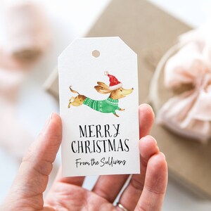 May include: White gift tag with a watercolor illustration of a dachshund wearing a green sweater and a red Santa hat. The text "Merry Christmas" and "From the Sullivans" is printed on the tag.