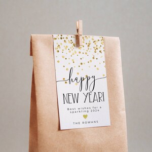 Puede incluir: Una etiqueta de regalo blanca con confeti dorado y el texto "happy new year!"  Los mejores deseos para un 2024 brillante. The Rowans.