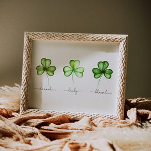 May include: A white framed print with three green shamrocks. The shamrocks are painted in watercolor and have the words "charmed", "lucky", and "blessed" written underneath them.