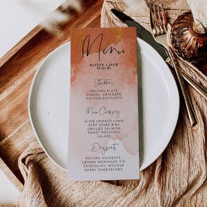 May include: A rectangular paper menu with a watercolor wash in shades of terracotta and cream. The text reads Menu, Alyssa + Jack 16.01.21, Starters, Main Courses, and Desserts, followed by a list of food items including lamb, salmon, and desserts.