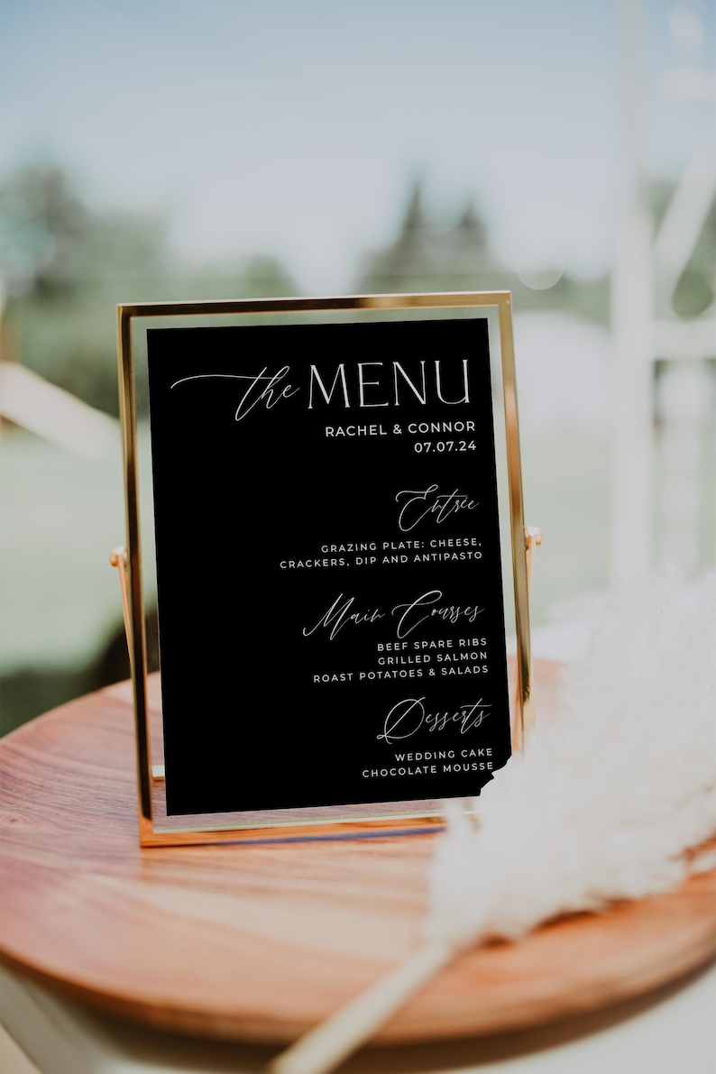 May include: A black framed menu board with gold trim, listing the menu for a wedding reception. The menu includes appetizers, main courses, and desserts. The text on the menu board reads "The Menu", "Rachel & Connor", "07.07.24", "Extre", "Grazing Plate: Cheese, Crackers, Dip and Antipasto", "Main Courses", "Beef Spare Ribs", "Grilled Salmon", "Roast Potatoes & Salads", "Desserts", "Wedding Cake", and "Chocolate Mousse".