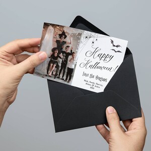 May include: A rectangular Halloween greeting card featuring a photograph of a family in costumes on the left and white space with black spiderweb, spider, and bat illustrations on the right. The text reads Happy Halloween! Love the Morgans, Nick, Sarah, Harvey, Rhien & Aiden 2022. The card is partially inserted into a black envelope.
