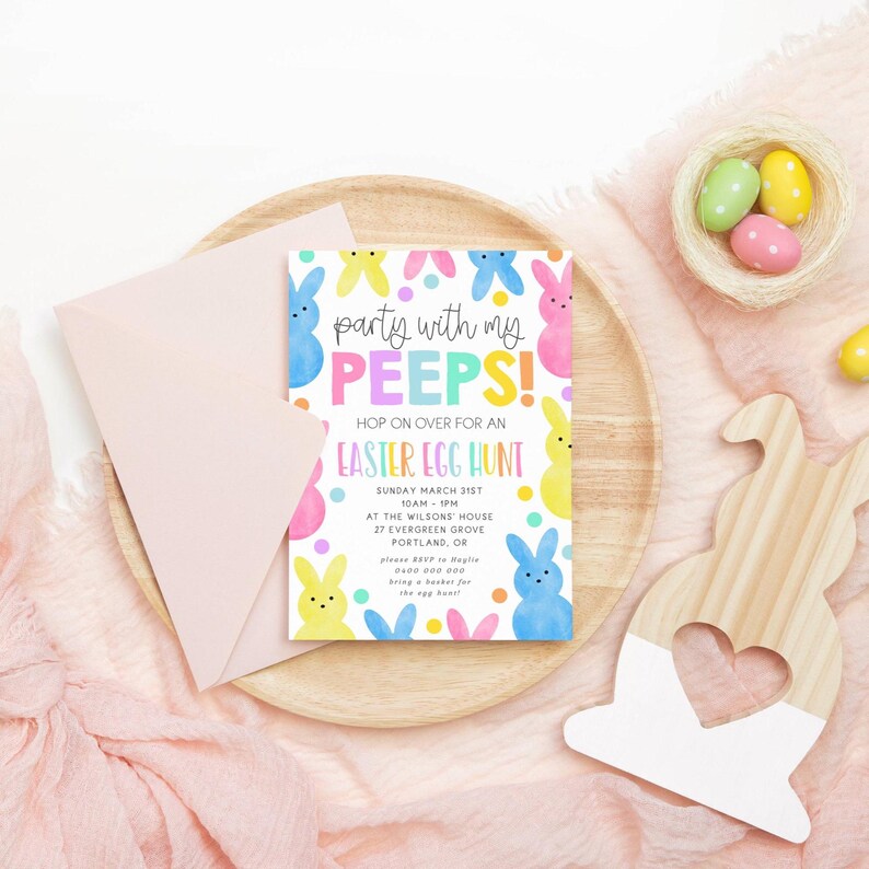 May include: An Easter-themed invitation with the text "Party with my PEEPS!" and "Easter Egg Hunt." The invitation is surrounded by colorful bunny illustrations and rests on a wooden tray with a pink envelope. A wooden bunny cutout and a nest with Easter eggs are also present.